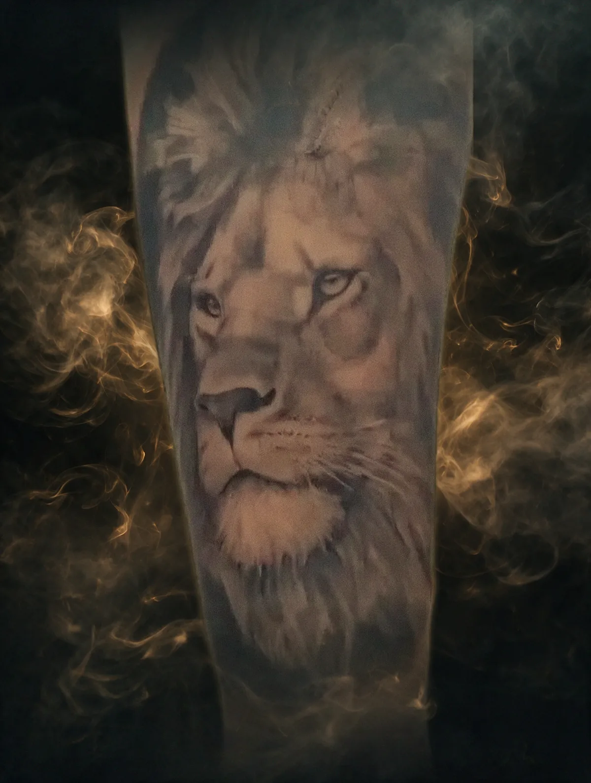 León tatuado en realismo black & grey sobre un antebrazo, emergiendo entre humo dorado