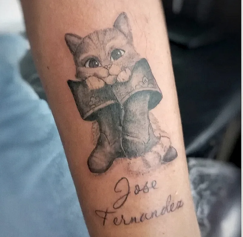 Gato con botas ilustrado con un nombre en lettering, tatuado en el antebrazo
